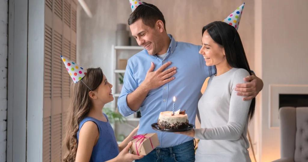 A happy family celebrating a birthday with party hats, a cake, and a gift. The father looks touched by the birthday wishes.
