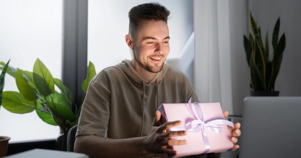 A happy man opening a promotional gift in Dubai, wrapped in pink with a ribbon, in a cozy home office setting.
