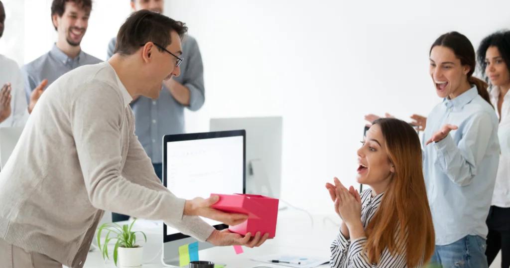 A man presenting a gift to a smiling woman at the office, surrounded by colleagues applauding. Perfect example of corporate gifting in the UK.