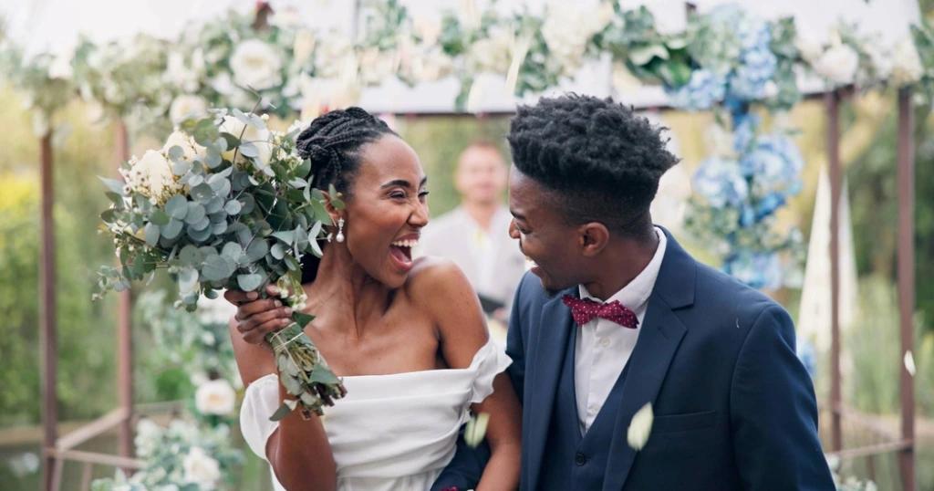A joyful bride and groom laugh together while the bride holds a vibrant wedding gift flower bouquet.